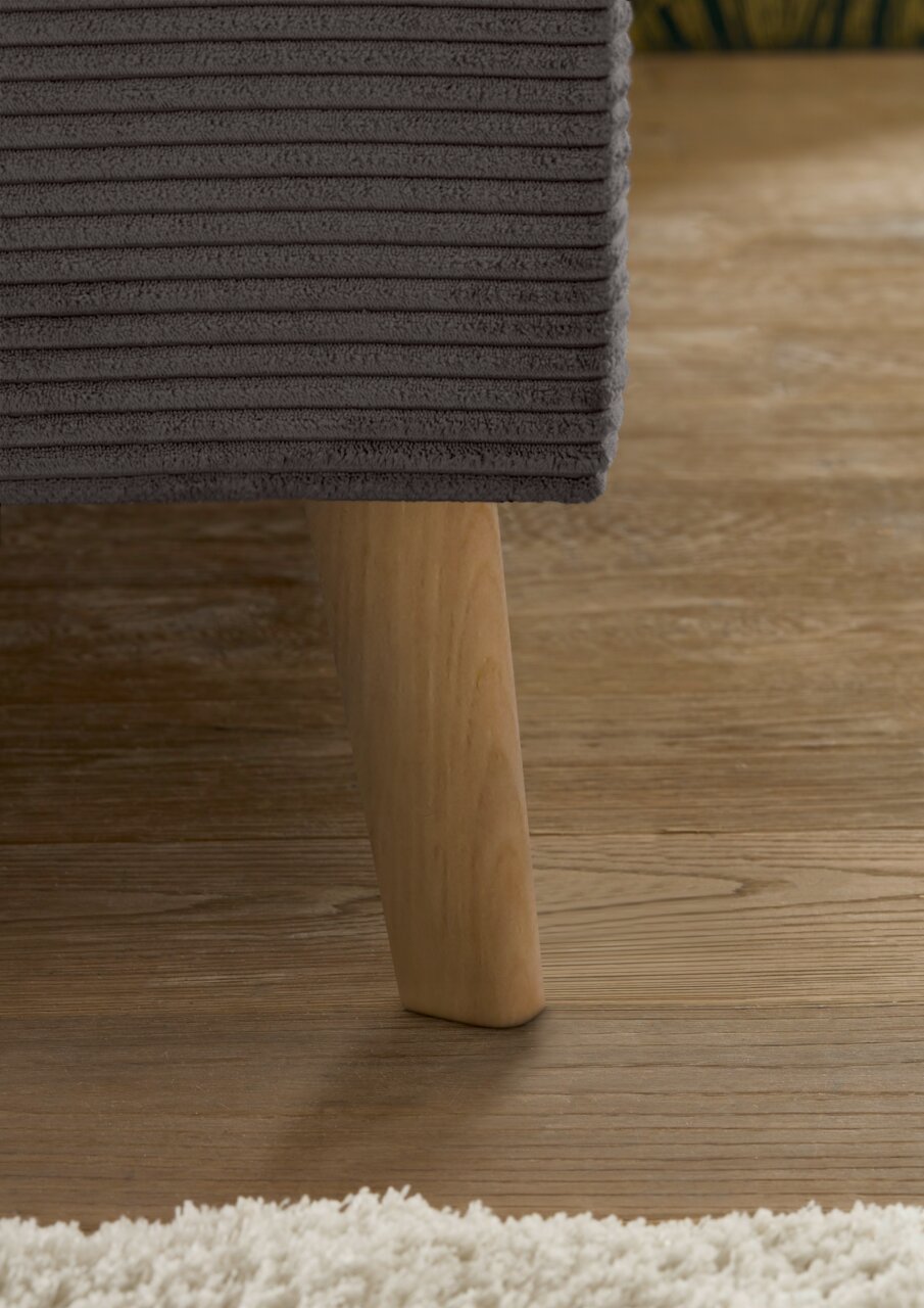 Close-up van een gestoffeerd bed met een donkergrijze, geribbelde stoffen bekleding en een houten poot op een houten vloer. Zijaanzicht.