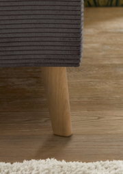 Close-up van een gestoffeerd bed met een donkergrijze, geribbelde stoffen bekleding en een houten poot op een houten vloer. Zijaanzicht.