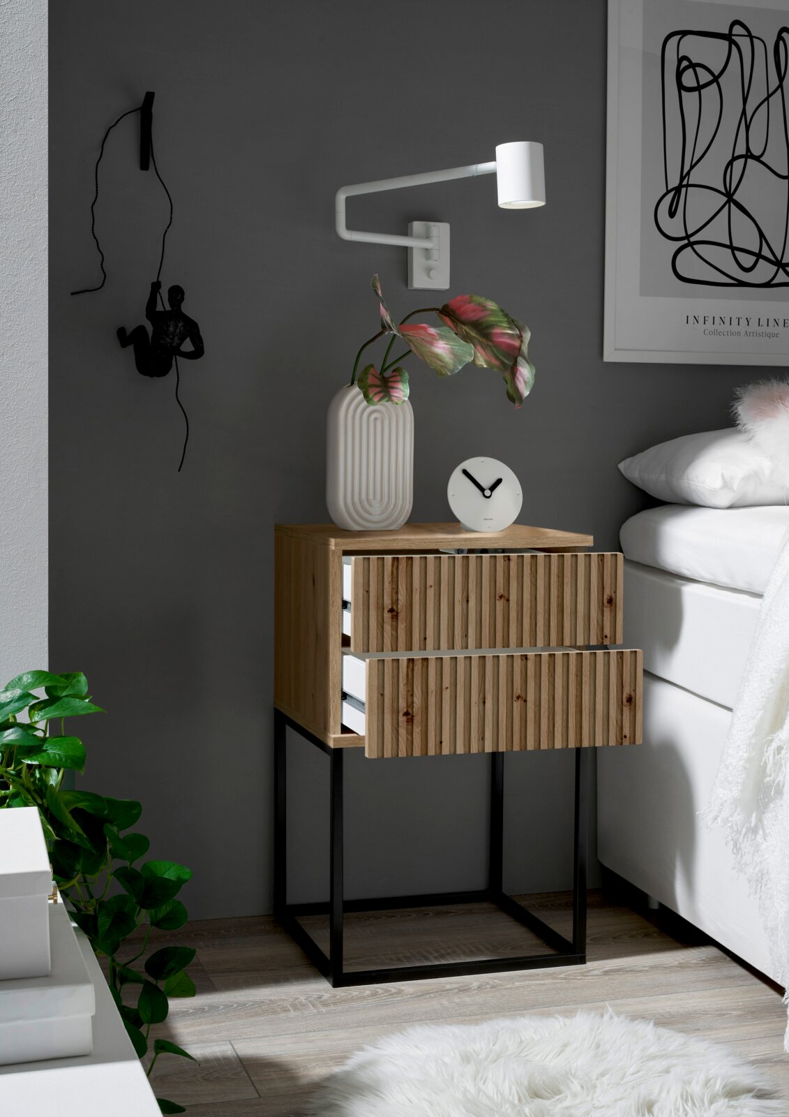 Nachtkommode aus Holz mit zwei Schubladen, auf einem schwarzen Metallgestell, seitliche Perspektive. Dekoriert mit einer weißen Vase, einer Uhr und einer Wandlampe.