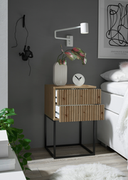 Nachtkommode aus Holz mit zwei Schubladen, auf einem schwarzen Metallgestell, seitliche Perspektive. Dekoriert mit einer weißen Vase, einer Uhr und einer Wandlampe.
