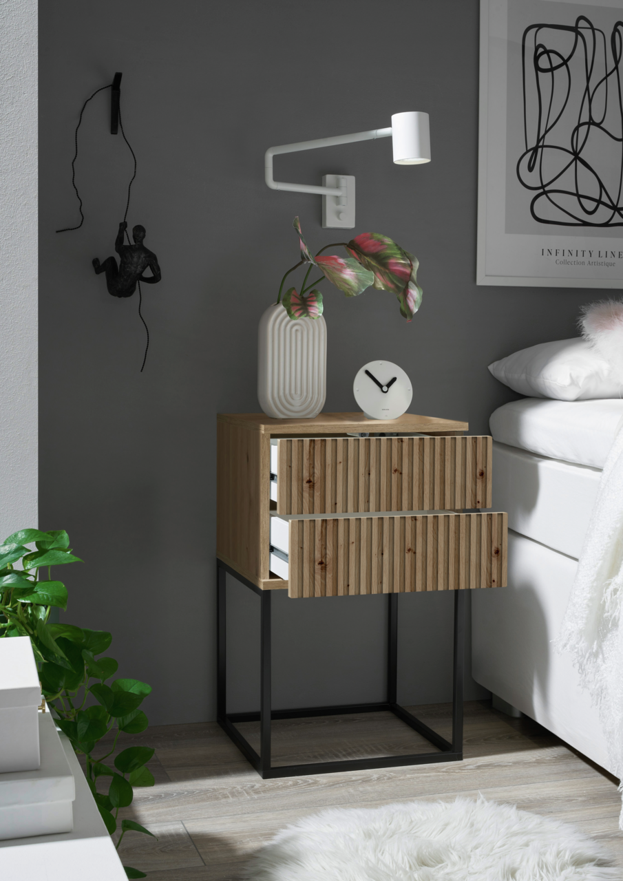 Nachtkommode aus Holz mit zwei Schubladen, auf einem schwarzen Metallgestell, seitliche Perspektive. Dekoriert mit einer weißen Vase, einer Uhr und einer Wandlampe.