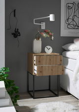 Nachtkommode aus Holz mit zwei Schubladen, auf einem schwarzen Metallgestell, seitliche Perspektive. Dekoriert mit einer weißen Vase, einer Uhr und einer Wandlampe.