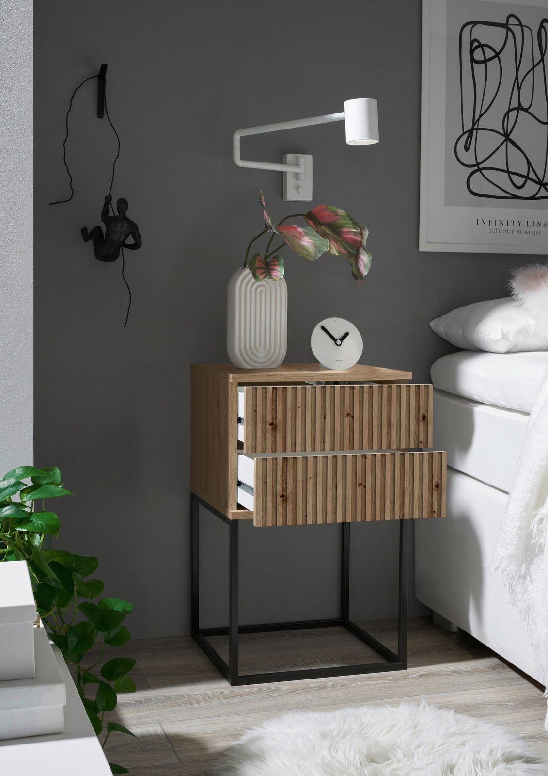 Nachtkommode aus Holz mit zwei Schubladen, auf einem schwarzen Metallgestell, seitliche Perspektive. Dekoriert mit einer weißen Vase, einer Uhr und einer Wandlampe.
