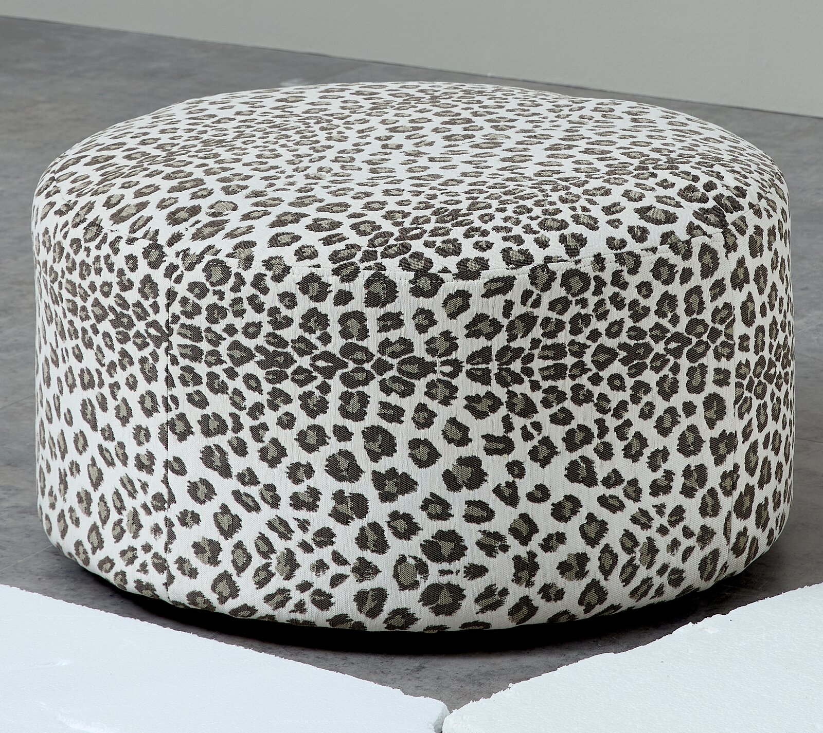 Runder, gepolsterter Pouf/Hocker mit Leopardenmuster in Weiß und Grau, freigestellt auf grauem Boden, Ansicht schräg von vorne leicht von oben