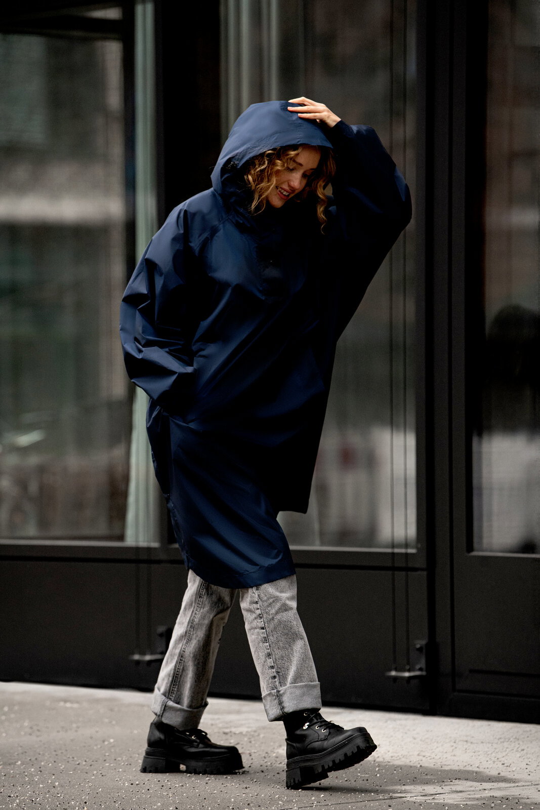 Person trägt einen dunkelblauen, oversized Regenponcho mit Kapuze und geht an einer Glasfassade entlang; Seitenansicht in voller Körperlänge.