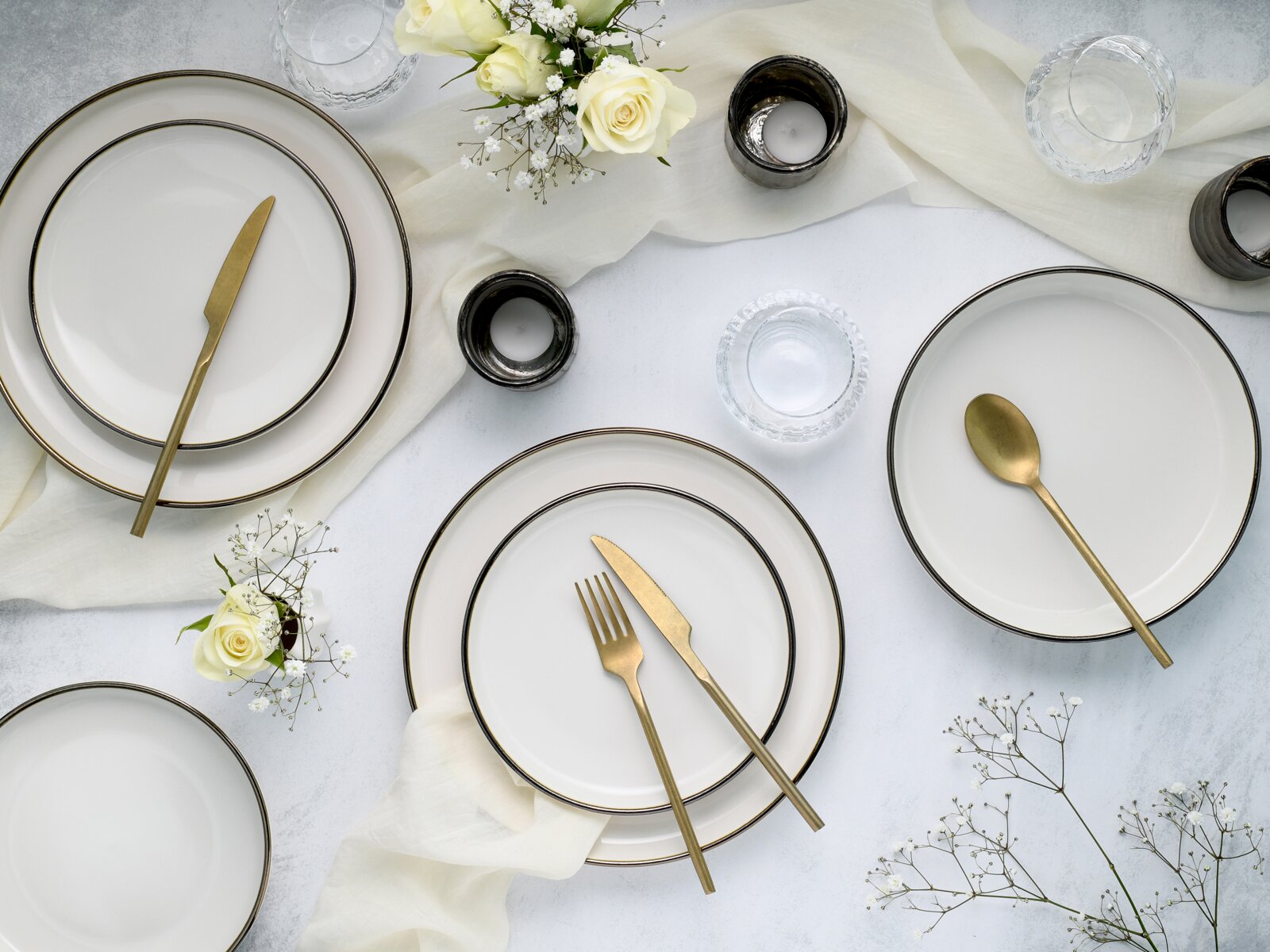 Draufsicht auf elegant gedeckten Tisch mit weißen Tellern mit schwarzem Rand, goldfarbenem Besteck, Kerzenhaltern, Wassergläsern und weißen Rosen auf hellem Stoff