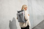Frau mit grauem urbanem Rolltop-Rucksack von der Seite fotografiert, vor einer Betonwand stehend.