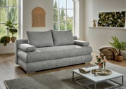 Graues Stoffsofa mit zwei großen Rückenkissen und breiten Armlehnen im hellen Wohnzimmer, perspektivisch in schräger Vorderansicht von links fotografiert.