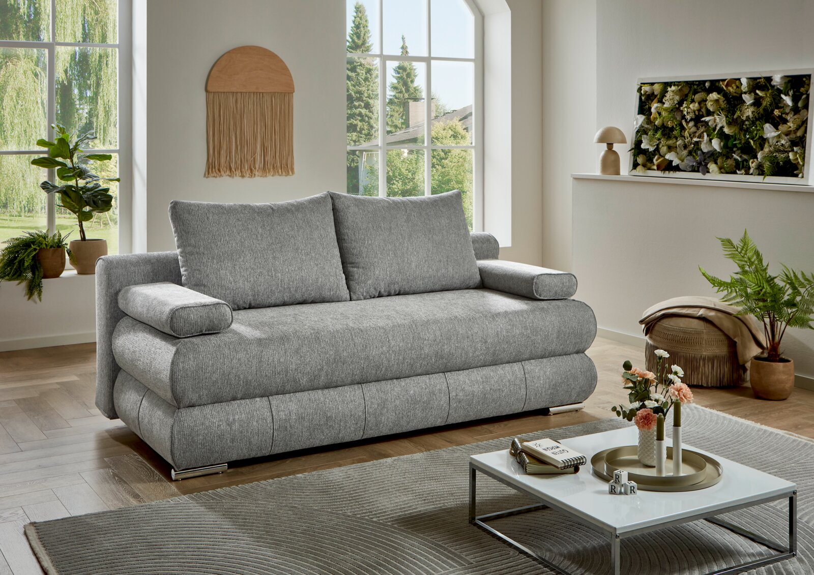 Graues Stoffsofa mit zwei großen Rückenkissen und breiten Armlehnen im hellen Wohnzimmer, perspektivisch in schräger Vorderansicht von links fotografiert.