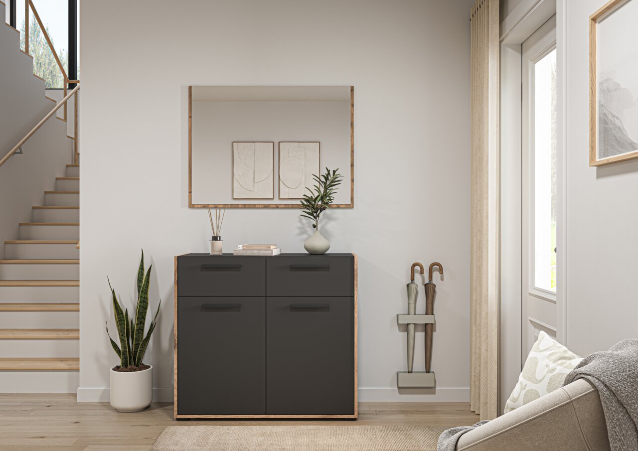 Frontale Ansicht einer modernen schwarzen Kommode mit Holzrahmen im Flur, dekoriert mit Vase und Duftstäbchen, darunter ein großer Wandspiegel; daneben Pflanzen, Treppe und Schirmständer.