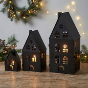 Drei schwarze Metall-Laternen in Hausform mit warmem Kerzenlicht, auf einem Holztisch vor weihnachtlicher Tannendeko und Lichterkette, fotografiert in leicht schräger Frontansicht auf Augenhöhe.
