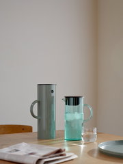 Zwei stilvolle Kannen auf einem Tisch, eine aus Metall und eine aus Glas, mit einem Glas Wasser und einer Zeitung im Vordergrund, seitliche Perspektive.