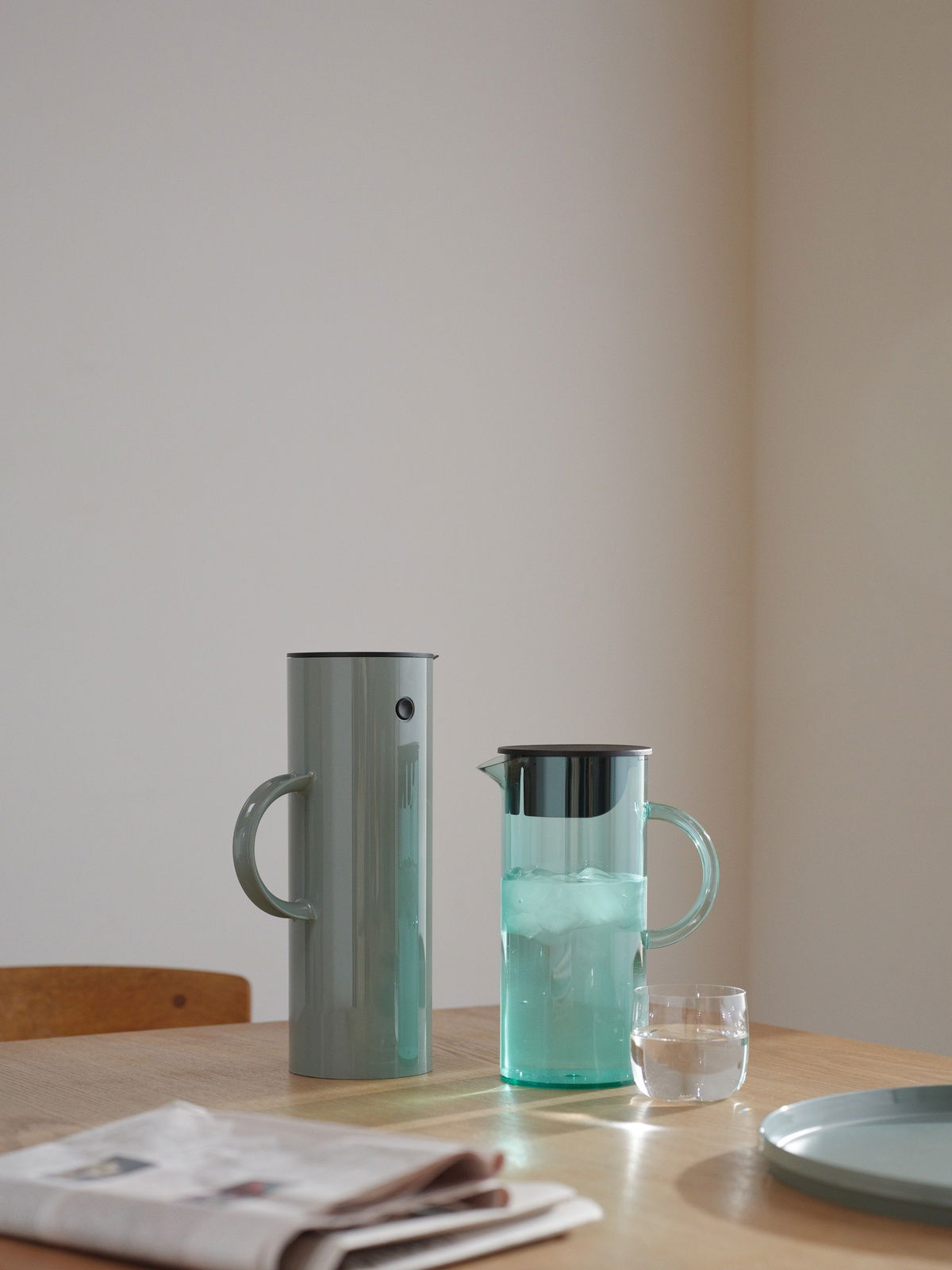 Zwei stilvolle Kannen auf einem Tisch, eine aus Metall und eine aus Glas, mit einem Glas Wasser und einer Zeitung im Vordergrund, seitliche Perspektive.
