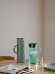 Zwei stilvolle Kannen auf einem Tisch, eine aus Metall und eine aus Glas, mit einem Glas Wasser und einer Zeitung im Vordergrund, seitliche Perspektive.