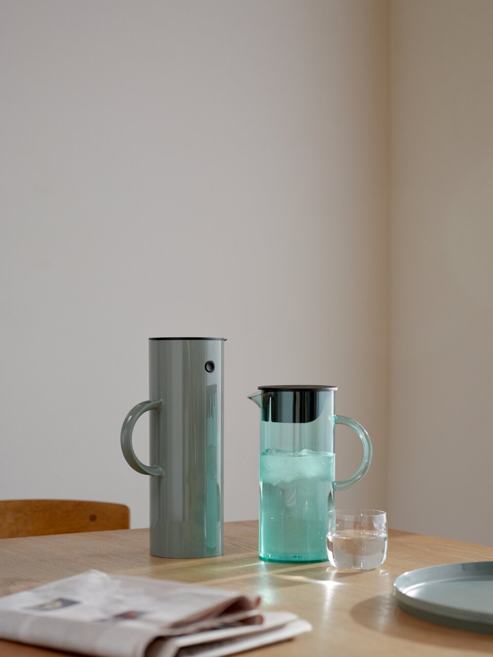 Zwei stilvolle Kannen auf einem Tisch, eine aus Metall und eine aus Glas, mit einem Glas Wasser und einer Zeitung im Vordergrund, seitliche Perspektive.