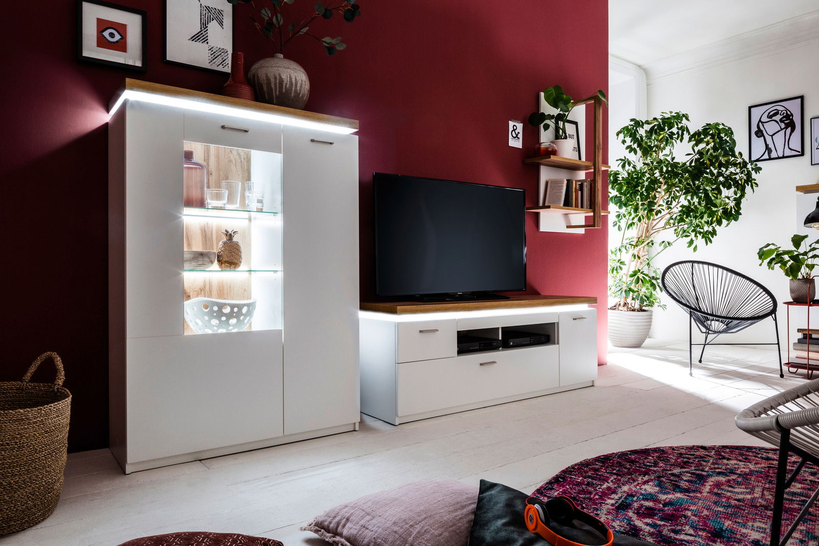 Wohnzimmeransicht mit modernem weißen Highboard und TV-Element vor einer roten Wand, dekoriert mit Pflanzen und Kunstwerken.