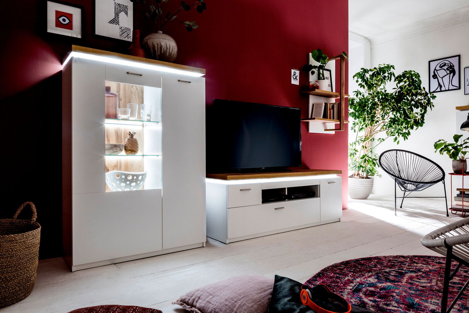 Wohnzimmeransicht mit modernem weißen Highboard und TV-Element vor einer roten Wand, dekoriert mit Pflanzen und Kunstwerken.