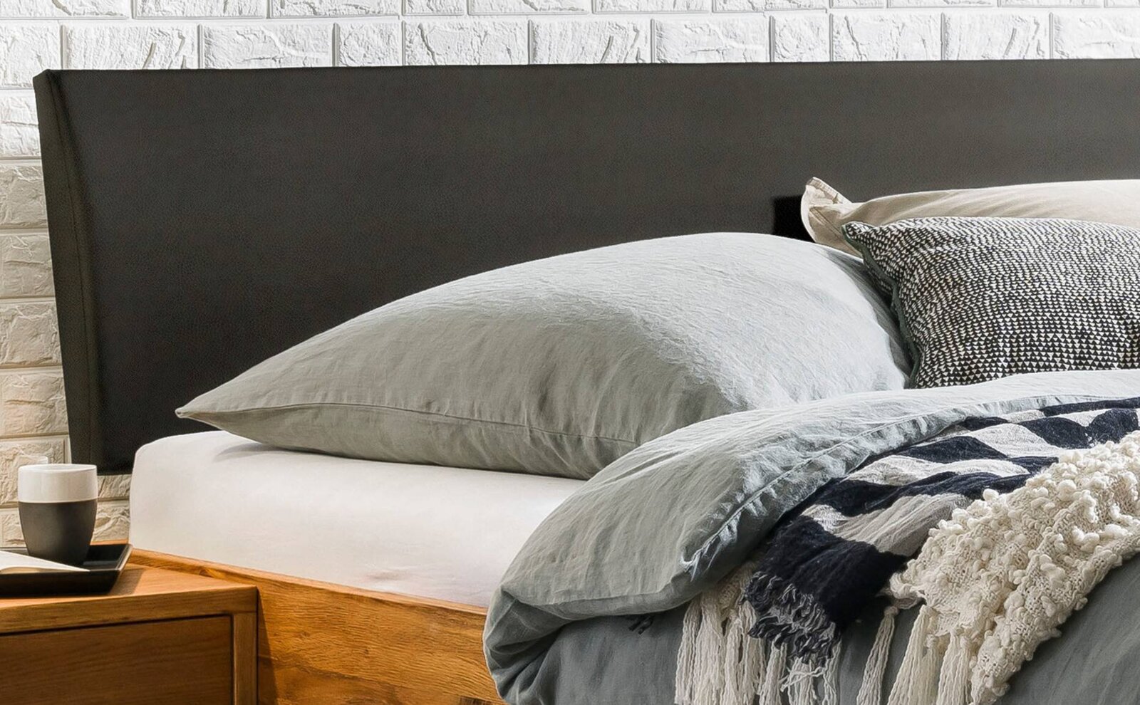 Modernes Bett mit gepolstertem, dunkelgrauem Kopfteil, grauer Bettwäsche und Kissen vor weißer Ziegelwand, aufgenommen in seitlicher Nahansicht aus leicht schräger Perspektive.