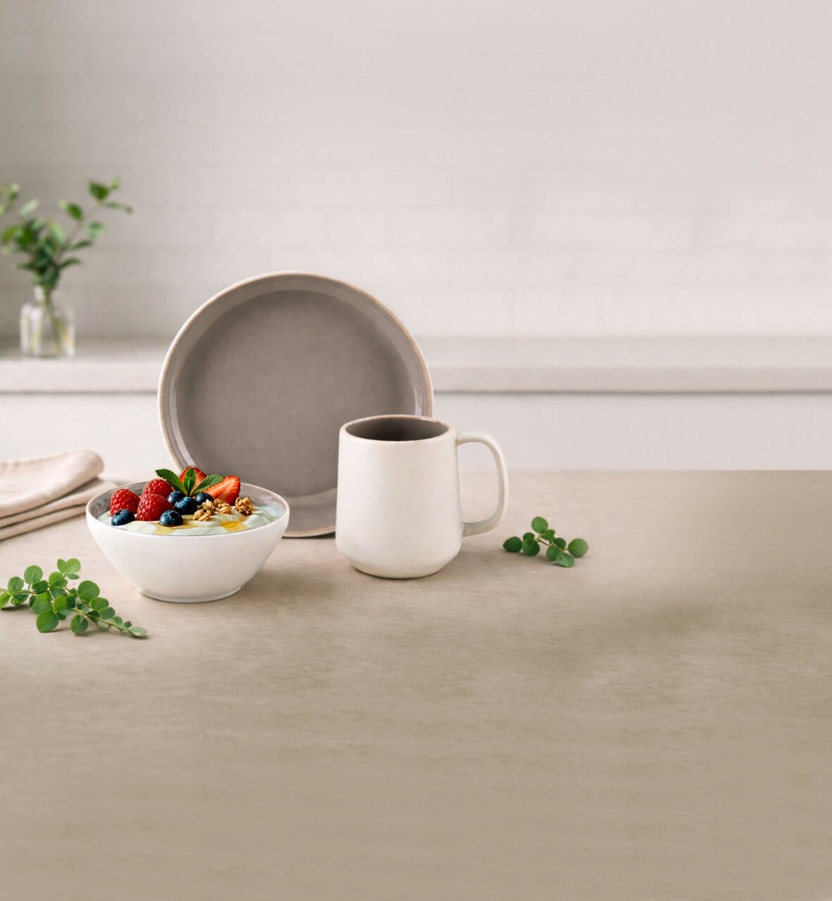 Minimalistisches Geschirr-Set auf hellem Tisch: grauer Teller im Hintergrund, weiße Kaffeetasse und Schale mit Joghurt, Beeren und Nüssen; Perspektive: frontale Tischhöhe mit leicht erhöhter Ansicht.