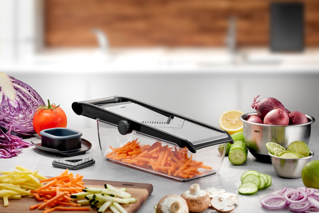 V-Hobel Julienne mit transparentem Auffangbehälter, gefüllt mit geschnittenen Karotten, umgeben von frischem Gemüse in einer modernen Küchenumgebung, seitliche Perspektive.