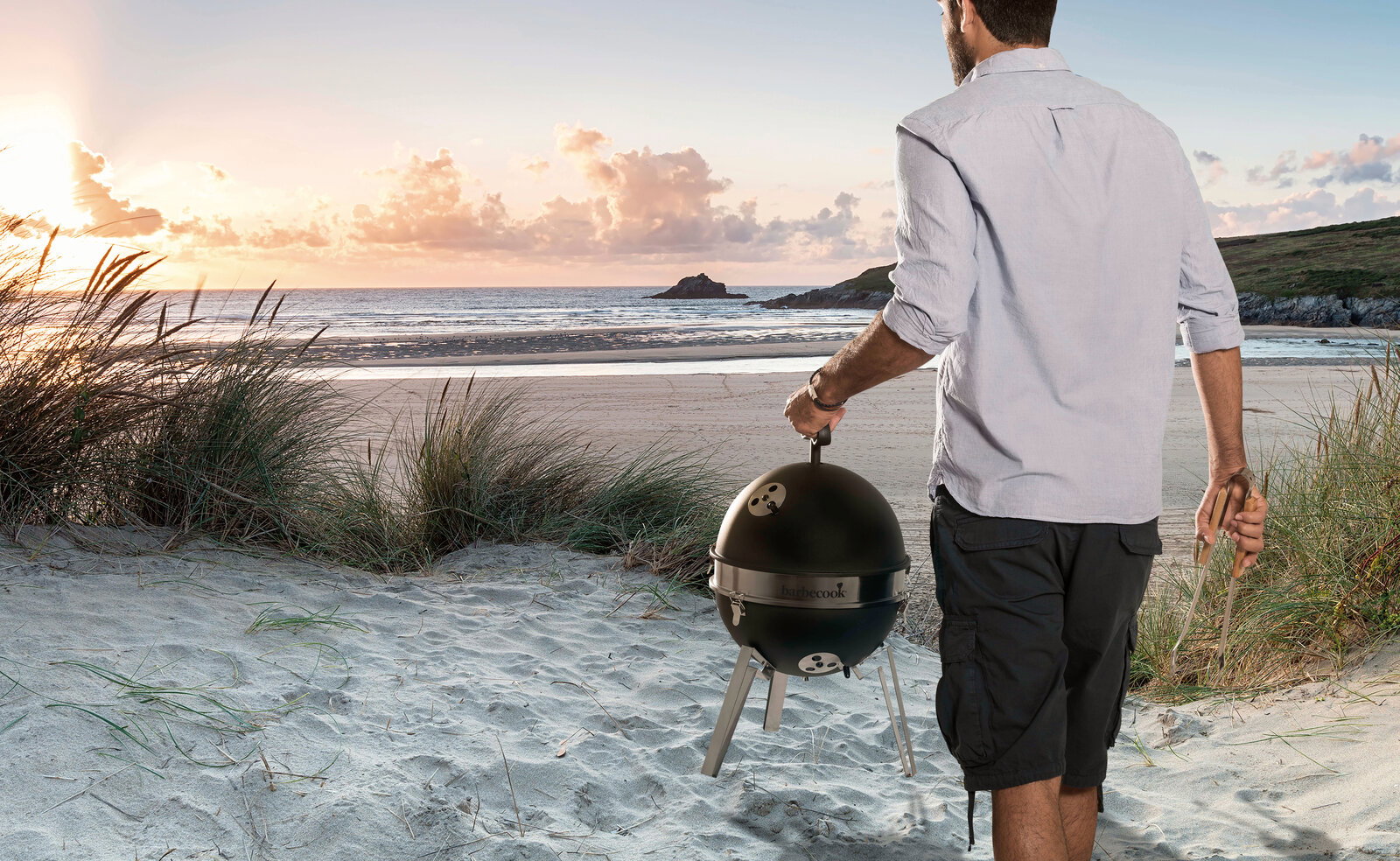 Barbecook BBQ om mee te nemen BILLY Mann von hinten in Stranddünen trägt einen kompakten schwarzen Kugelgrill mit Standbeinen Richtung Meer bei Sonnenuntergang, Perspektive: Rückansicht/leicht seitlich.