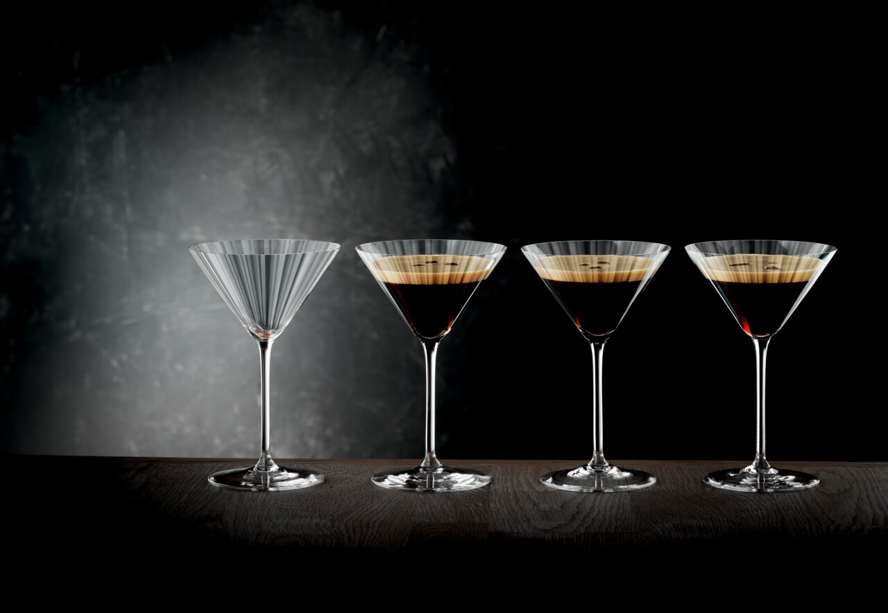 Spiegelau Martini-Set 4 tlg. LIFESTYLE Vier Martini-Gläser, eines leer und drei mit dunklem Getränk gefüllt, auf einer Tischoberfläche vor einem dunklen Hintergrund, seitliche Perspektive.