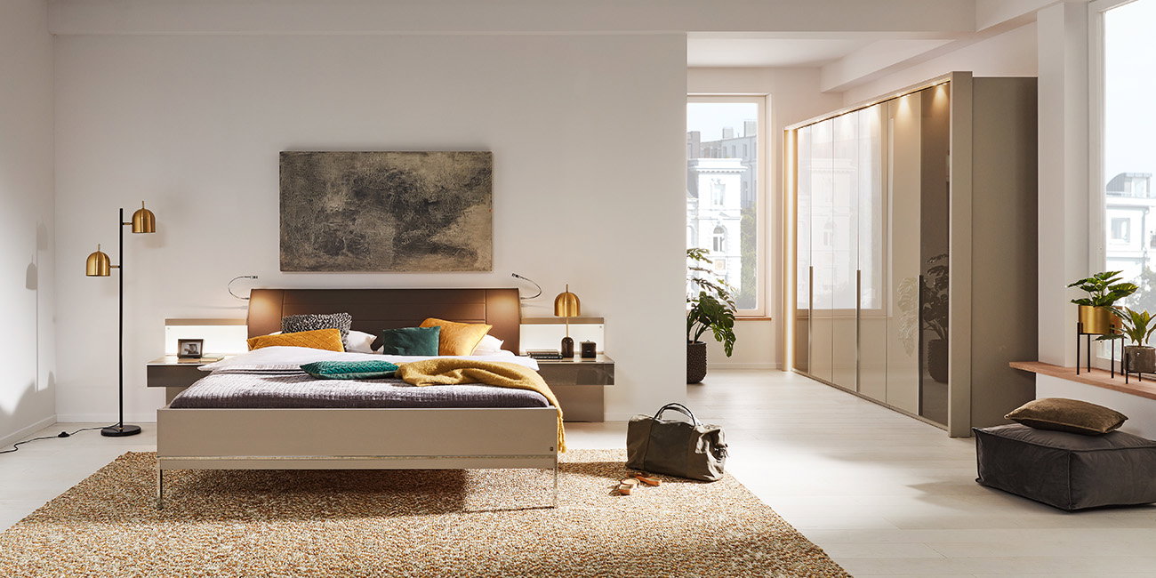 Das Bild zeigt ein modernes Schlafzimmer. In der Mitte steht ein großes Bett mit einer gemütlichen Decke und vielen Kissen in verschiedenen Farben. Links und rechts neben dem Bett sind kleine Nachttische mit Lampen. Im Hintergrund steht ein großer Kleider