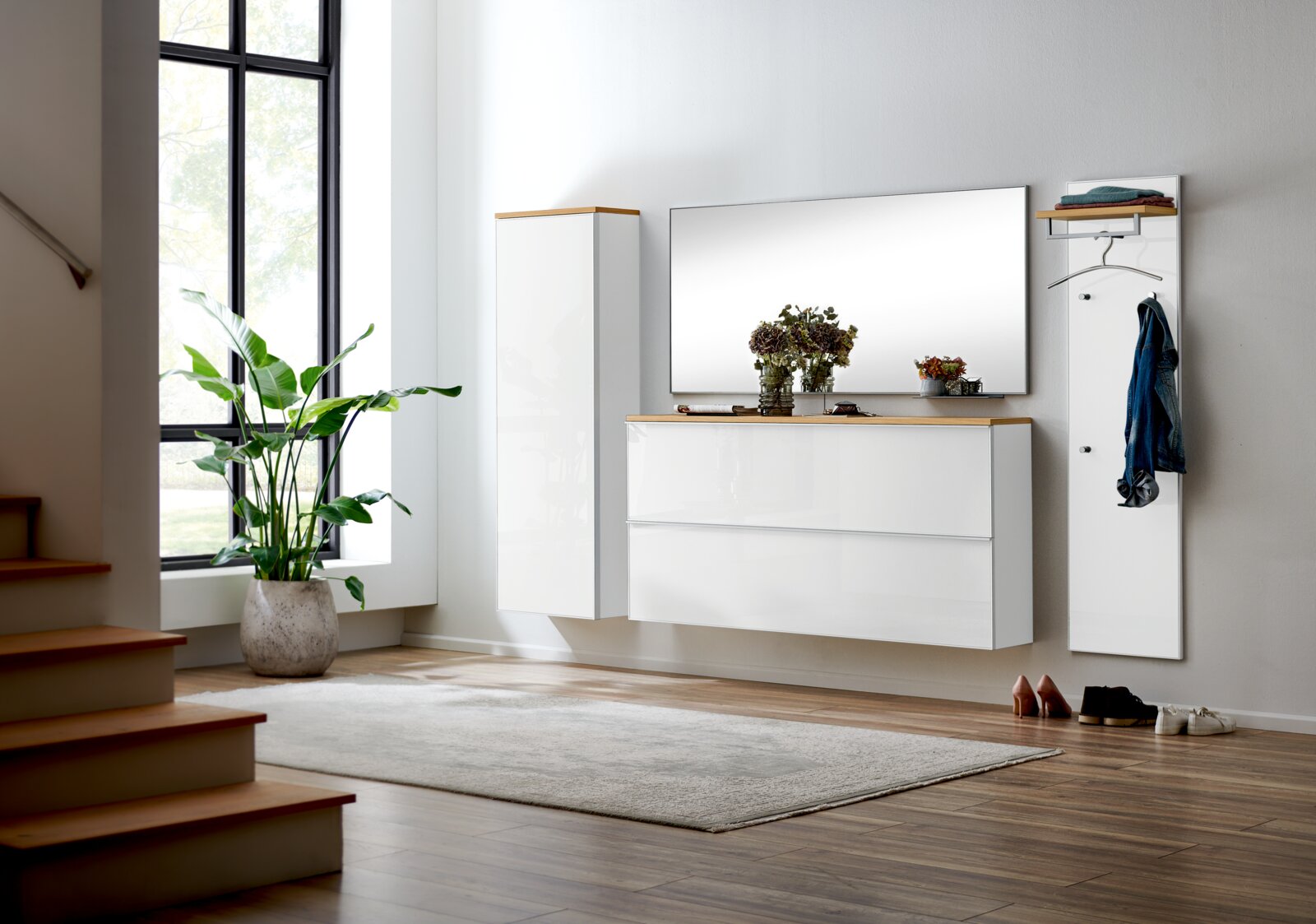 Modernes, wandhängendes Flurmöbel-Set in Weiß Hochglanz mit Holzablagen, großem Spiegel, Hängeschrank und Garderobenpaneel mit Haken und Kleiderbügel, frontal in leichter Schrägansicht von links im hellen Eingangsbereich
