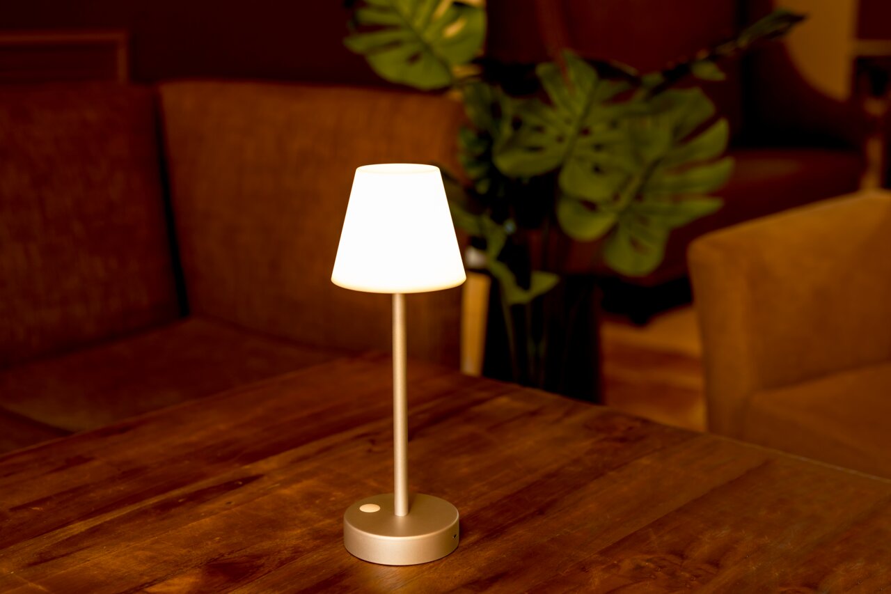Elegante Akku-Tischleuchte LOLA slim auf einem Holztisch, seitliche Perspektive, mit warmem Lichtschein in gemütlicher Umgebung.