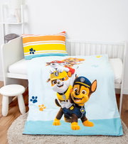 Renforcé-Bettwäsche mit buntem Paw Patrol-Motiv, bestehend aus einem Kissenbezug mit Streifenmuster und einer Bettdecke mit fröhlichen Hundemotiven, auf einem Kinderbett von schräg oben fotografiert.