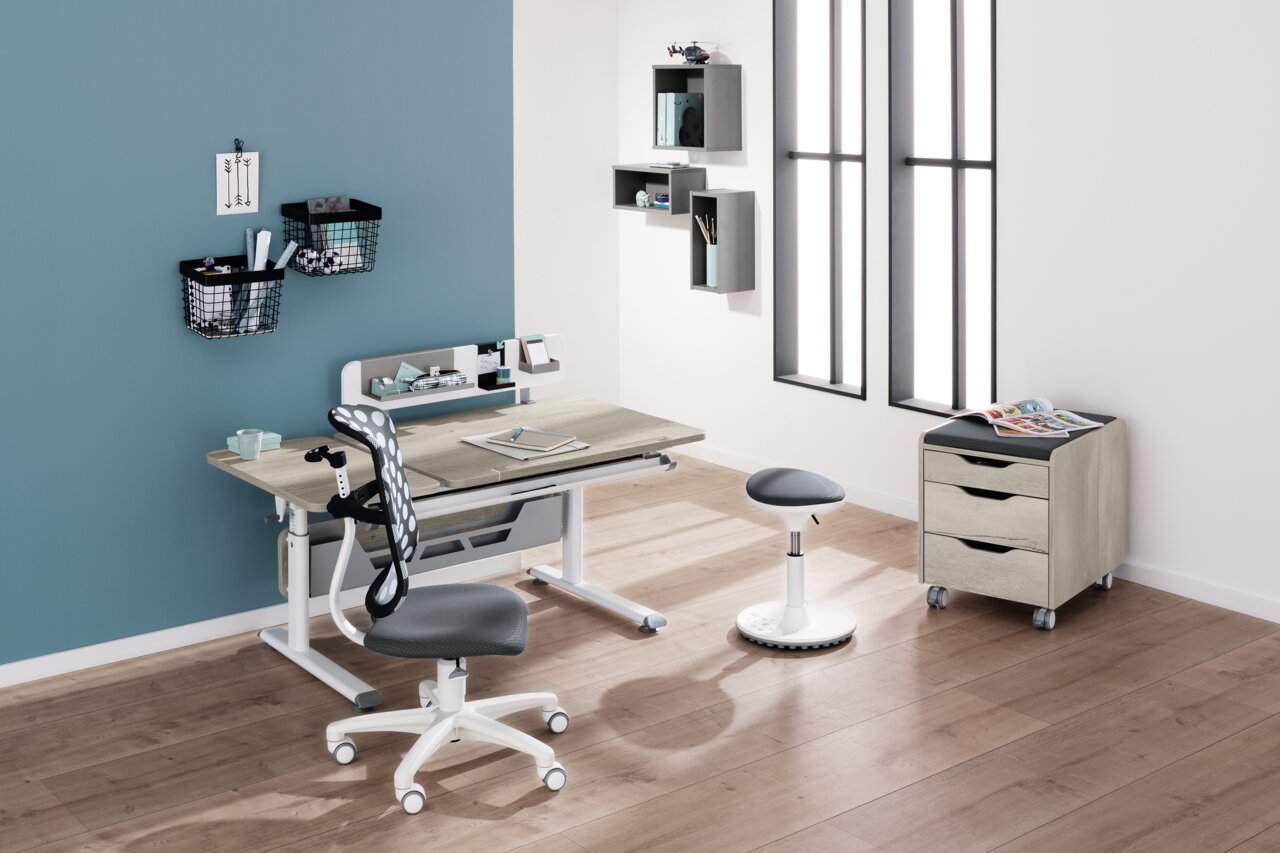 Moderner Schreibtisch mit ergonomischem Stuhl und Hocker in einem stilvollen, hellen Raum mit Holzboden, aus der Perspektive von vorne links betrachtet.