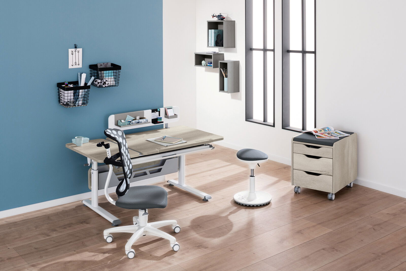 Moderner Schreibtisch mit ergonomischem Stuhl und Hocker in einem stilvollen, hellen Raum mit Holzboden, aus der Perspektive von vorne links betrachtet.