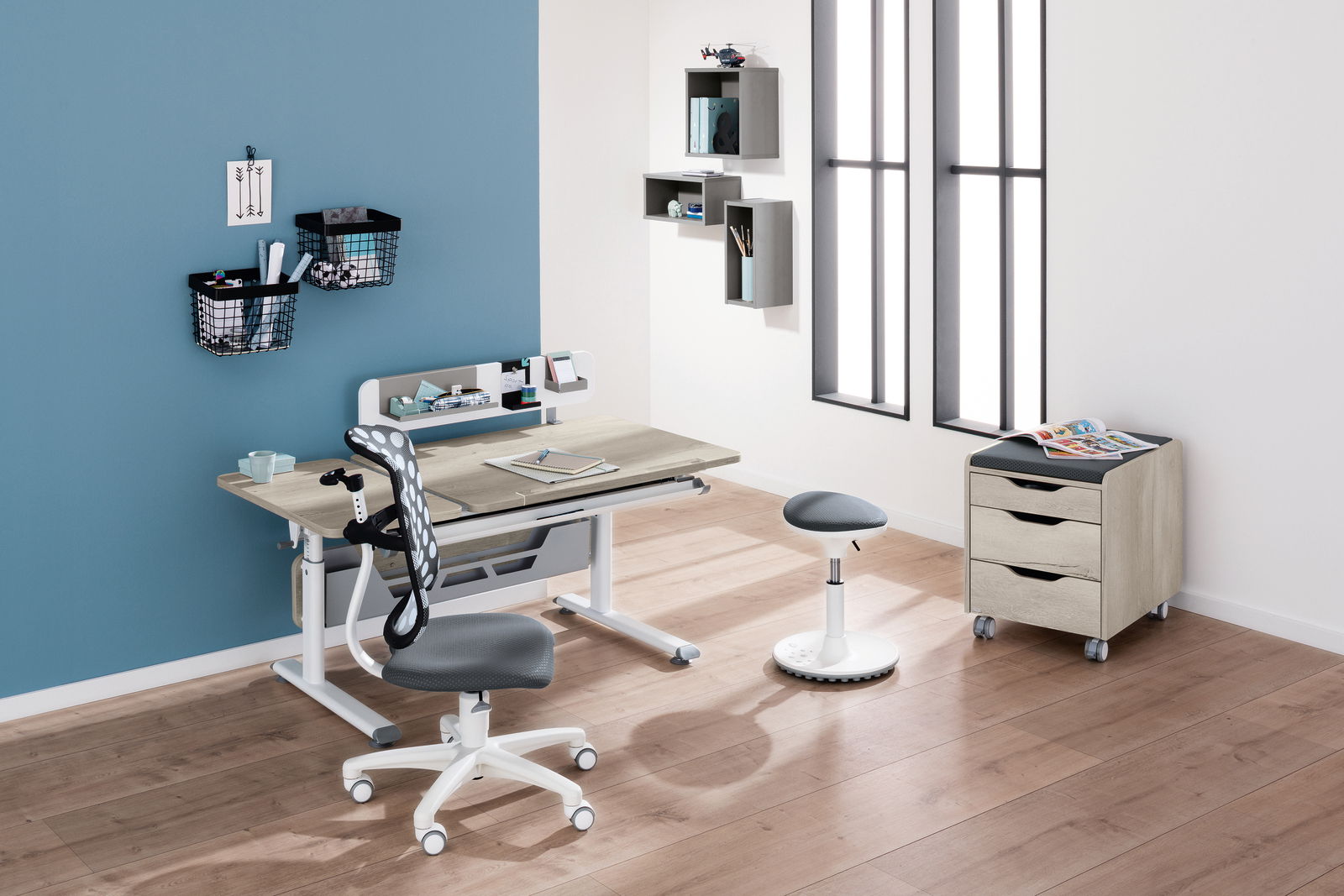Moderner Schreibtisch mit ergonomischem Stuhl und Hocker in einem stilvollen, hellen Raum mit Holzboden, aus der Perspektive von vorne links betrachtet.