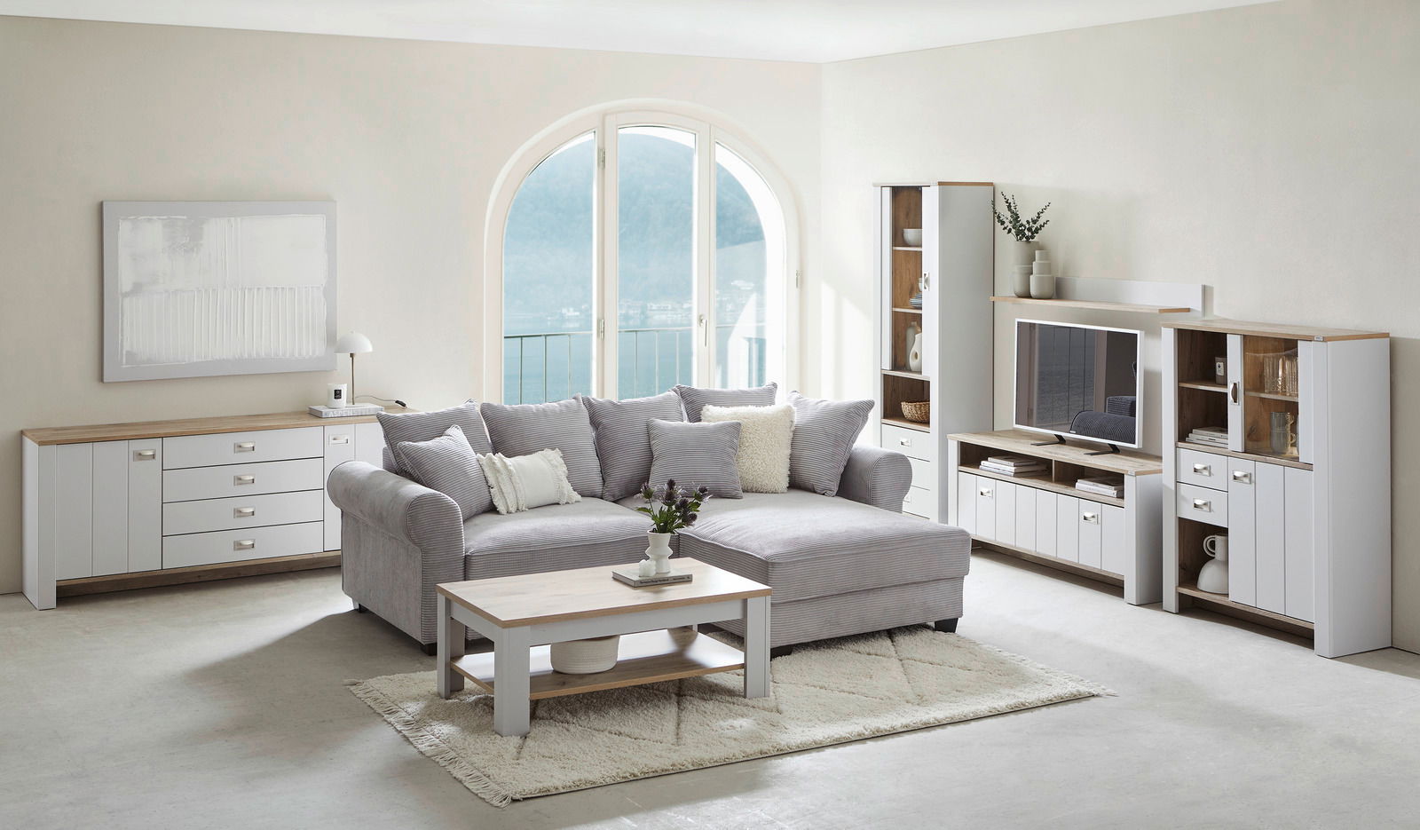 Wohnzimmer mit grauem Ecksofa, Couchtisch aus Holz und weißem Sideboard, Blick auf große Fenster mit Seeblick.