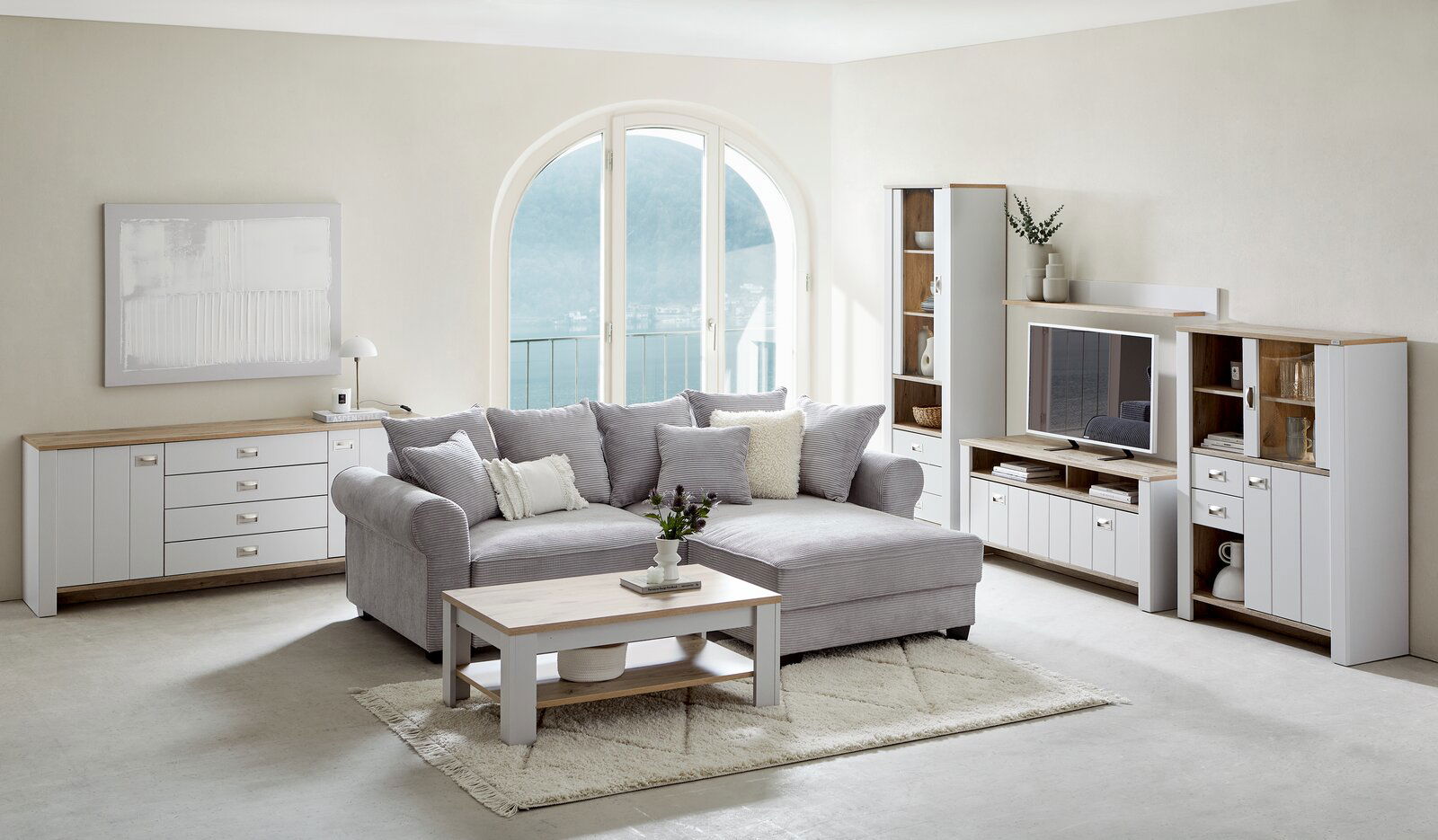 Wohnzimmer mit grauem Ecksofa, Couchtisch aus Holz und weißem Sideboard, Blick auf große Fenster mit Seeblick.
