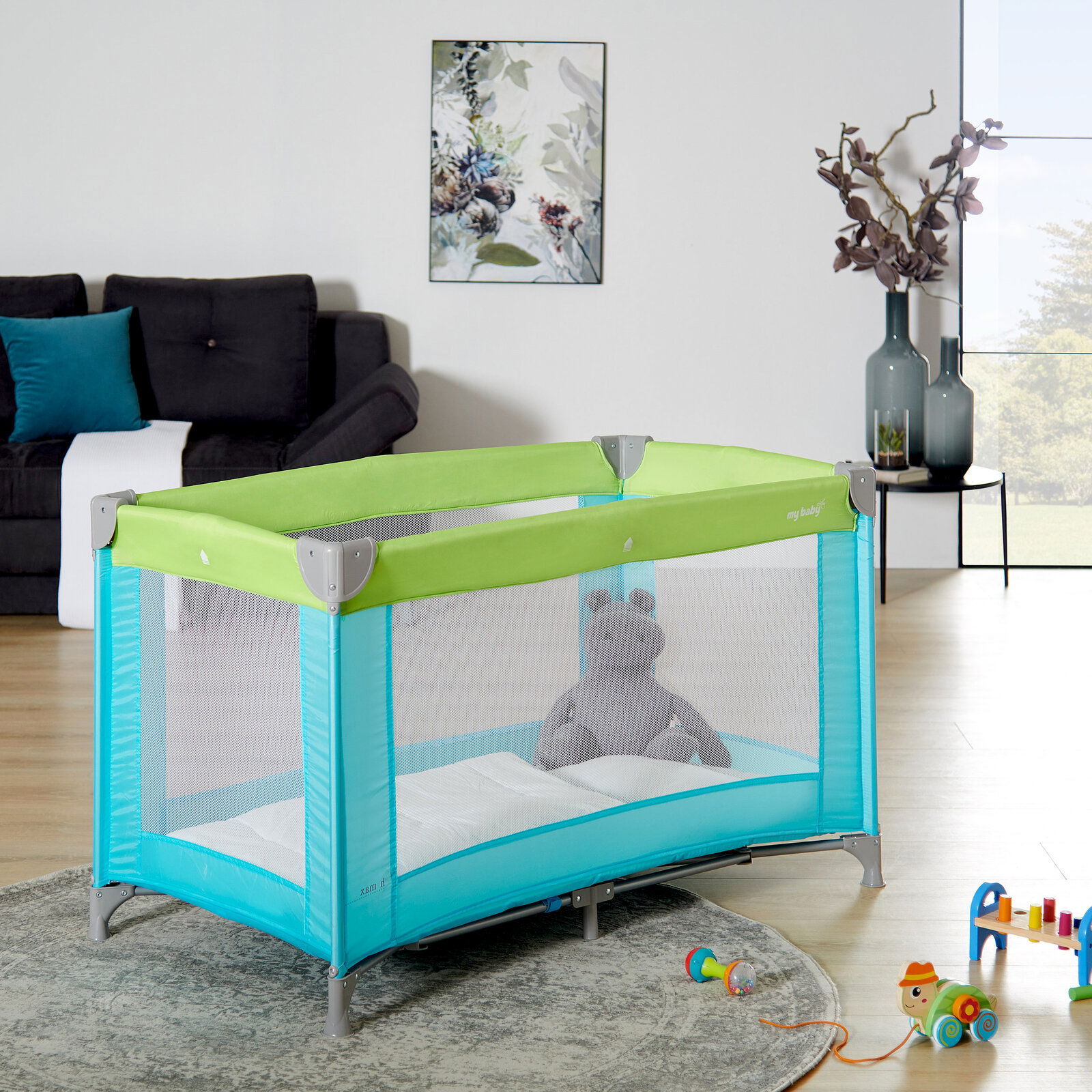 Reisebett in einem modernen Wohnzimmer, seitliche Perspektive, mit grünem und türkisfarbenem Design, gefüllt mit einem Plüschtier.