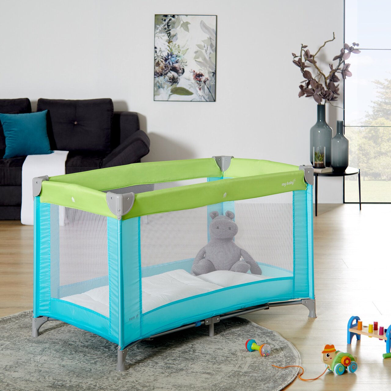 MyBaby Reisbedje Reisbed in een moderne woonkamer, gezien vanaf de zijkant, met een groen en turquoise patroon, met daarin een knuffel.