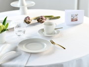 Hell gedeckter Tisch mit weißem Porzellanteller und -tasse, Wasserglas, goldfarbenem Besteck, Artischocken und Blumendeko auf weißer Tischdecke, fotografiert aus leicht erhöhter schräger Perspektive von vorn.