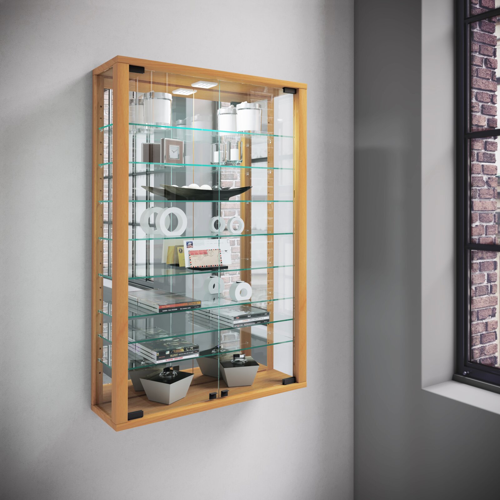 Wandhängende Glasvitrine mit hellem Holzrahmen und mehreren Glasböden, dekorativ bestückt, in schräger Frontansicht von links vor einer grauen Wand neben einem Fenster