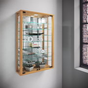 Wandhängende Glasvitrine mit hellem Holzrahmen und mehreren Glasböden, dekorativ bestückt, in schräger Frontansicht von links vor einer grauen Wand neben einem Fenster