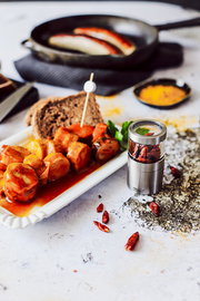 Mini Chili- und Gewürzschneider aus Edelstahl, gefüllt mit getrockneten Chilis, auf einem Tisch mit Gewürzen und einem Teller mit Wurstscheiben. Perspektive von vorne.