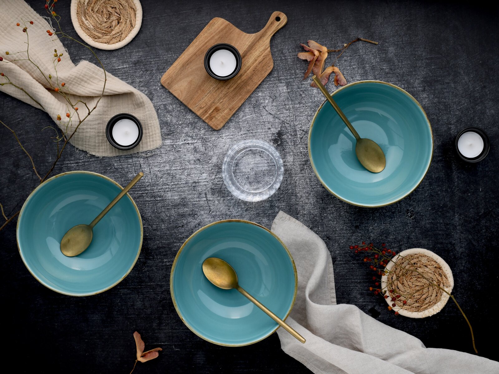 Drei türkisfarbene Keramikschalen mit goldfarbenen Löffeln auf dunkler Tischplatte, dekoriert mit Leinentüchern, Kerzen und Holzbrett – Draufsicht (Flatlay).