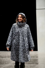 Frontansicht: Frau trägt einen grauen Regenmantel/Parka mit Kapuze im Leopardenmuster, Outdoor bei nassem Wetter vor dunklem Hintergrund.