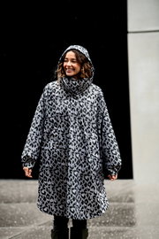 Frontansicht: Frau trägt einen grauen Regenmantel/Parka mit Kapuze im Leopardenmuster, Outdoor bei nassem Wetter vor dunklem Hintergrund.