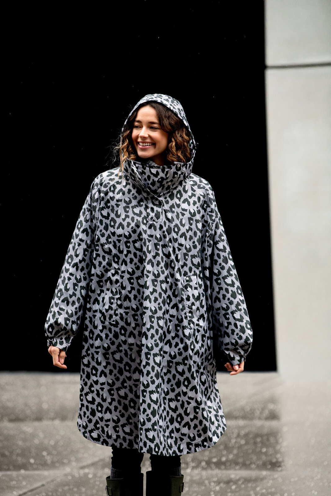Frontansicht: Frau trägt einen grauen Regenmantel/Parka mit Kapuze im Leopardenmuster, Outdoor bei nassem Wetter vor dunklem Hintergrund.