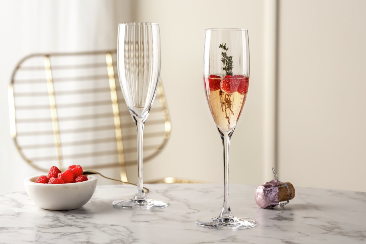 Zwei elegante Sektgläser POESIA 250ml auf einem Marmortisch, eines gefüllt mit Sekt, Himbeeren und einem Thymianzweig, im Hintergrund ein goldener Stuhl. Perspektive: seitlich.