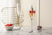 Zwei elegante Sektgläser POESIA 250ml auf einem Marmortisch, eines gefüllt mit Sekt, Himbeeren und einem Thymianzweig, im Hintergrund ein goldener Stuhl. Perspektive: seitlich.