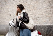 Een jonge vrouw, gezien vanaf de zijkant, draagt een crèmekleurige gewatteerde schoudertas en staat naast een witte scooter voor een lichte stenen muur; perspectief: zijaanzicht op heup-/borsthoogte.
