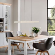 Moderner Esstisch mit weißem Gestell und heller Holzplatte, umgeben von gepolsterten Stühlen, im Esszimmer aus seitlicher Perspektive mit Blick auf den Tisch und die Hängelampe.
