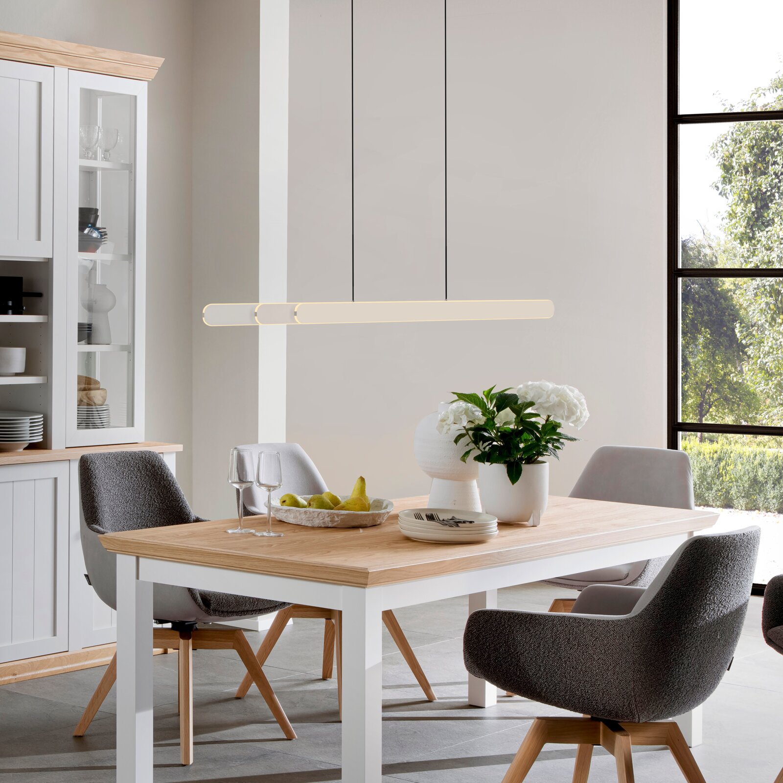 Moderner Esstisch mit weißem Gestell und heller Holzplatte, umgeben von gepolsterten Stühlen, im Esszimmer aus seitlicher Perspektive mit Blick auf den Tisch und die Hängelampe.